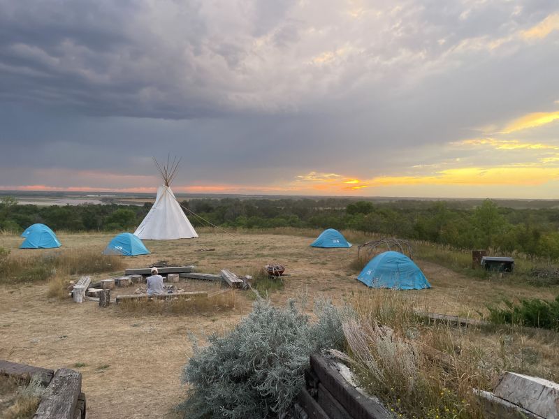 Campground in a grassy plain with multiple small blue camping tents and a large white tipi in the middle.