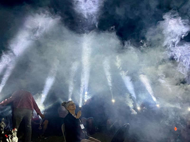 People sitting in a circle using torches pointed up at the sky to create a tipi shape using light.