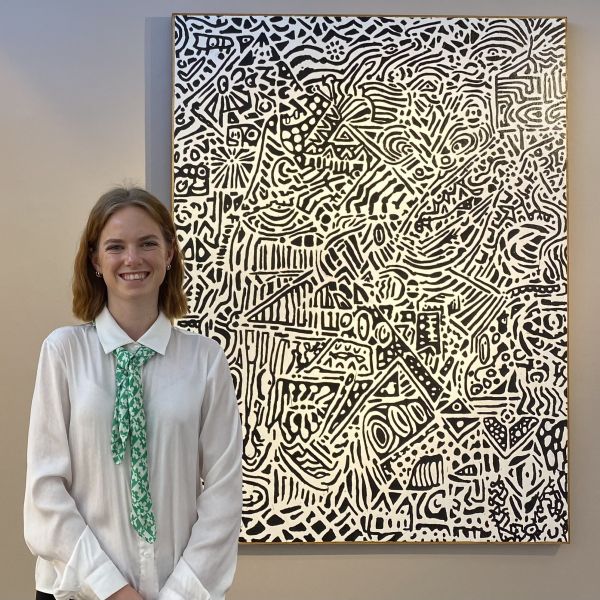 Ella stands in front of a white and black abstract painting of Ben Kenning's. She is dressed in a white button up and green tie, and beams at the camera. She has a short red bob.