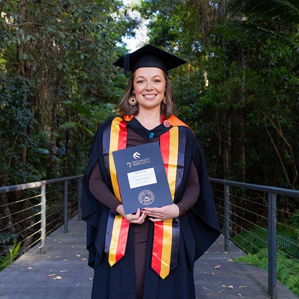 Kiah O'Toole stands wearing her graduation cap, gown, and Indigenous sash proudly holding her degree. Kicking goals in Nursing.
