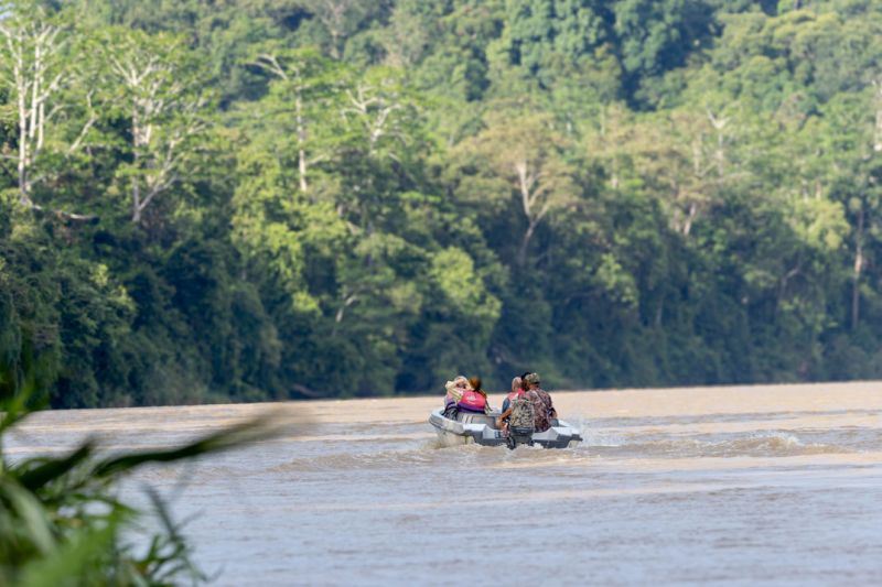 People wearing life jackets traveling down a river in a small motorboat, surrounded by lush forest.