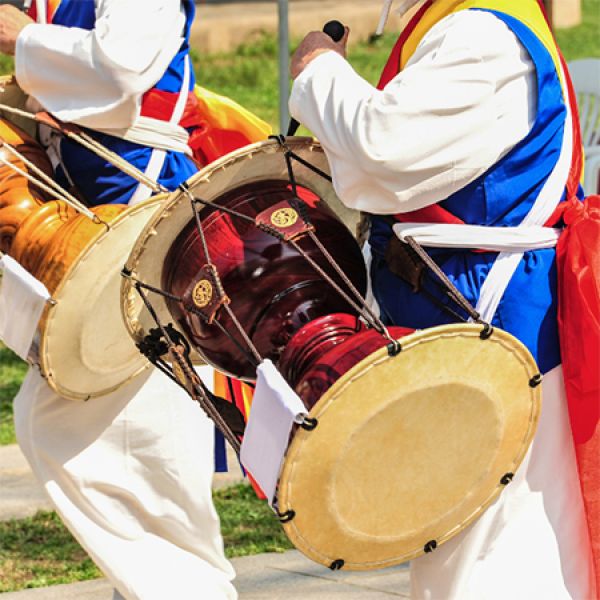 Two individuals in traditional Korean clothing play large double-headed drums outdoors.