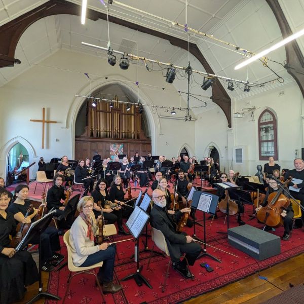 A large orchestra is seated in a church-like venue with high ceilings, an organ, and a cross on the wall.