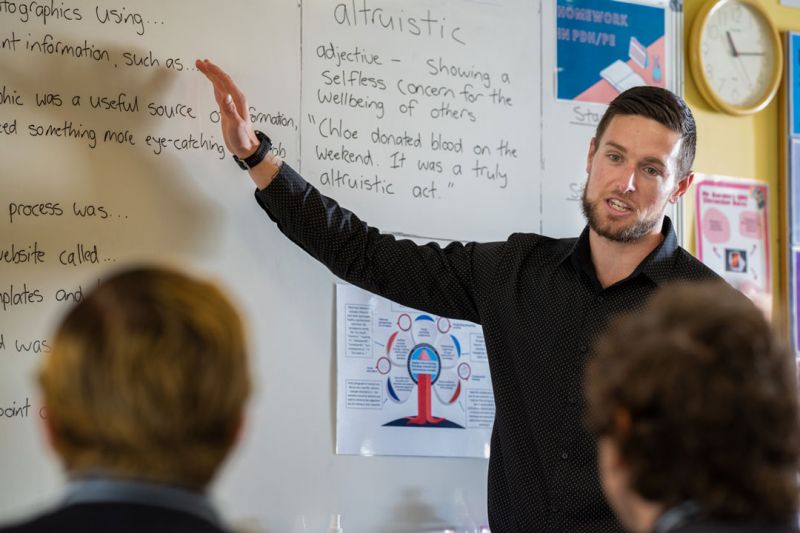 Josh, dressed in a long-sleeve black shirt, stands at the front of a classroom pointing to a whiteboard covered in notes as he explains a lesson to his students.
