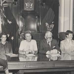 Black and white photo of four people at a stately desk with flags behind and the word "Newcastle"
