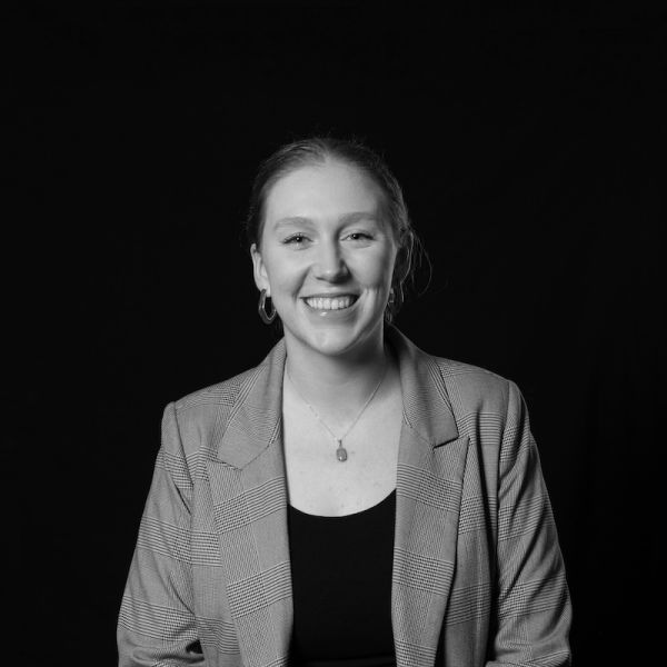 Smiling woman in a checkered blazer and hoop earrings against a dark backdrop. Her expression conveys friendliness and confidence. Black and white.