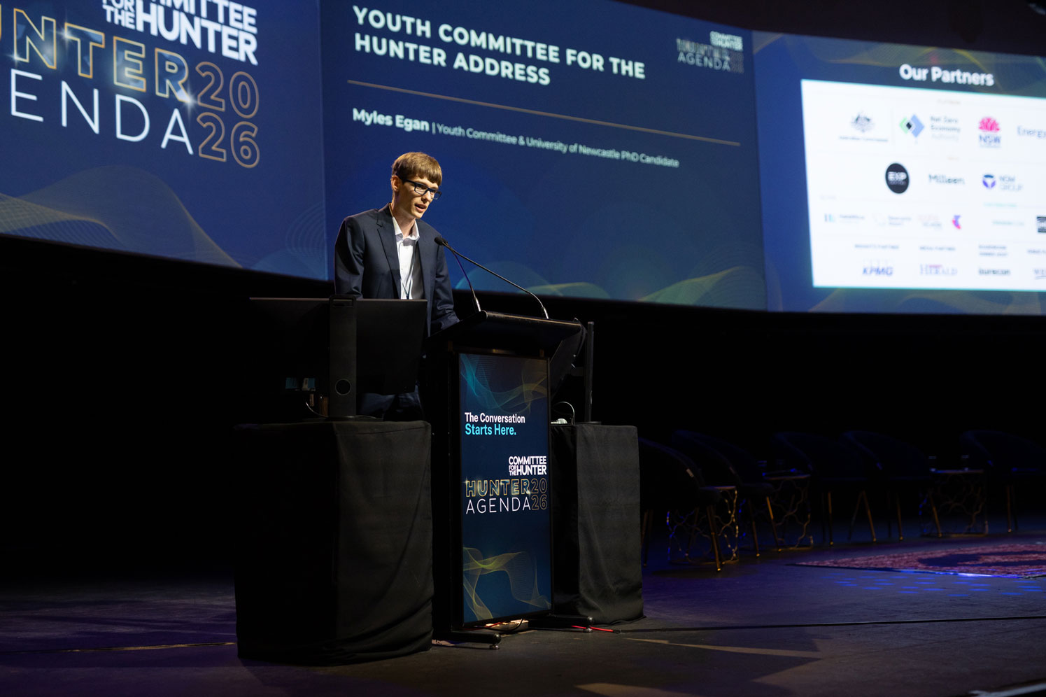 Myles Egan stands at a lectern on a stage, speaking into a microphone, in front of screens displaying event content