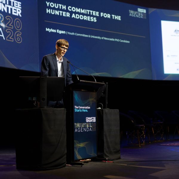 Myles Egan stands at a lectern on a stage, speaking into a microphone, in front of screens displaying event content. University PhD Candidate Amplifies Youth Voice in Regional Transition Talks.