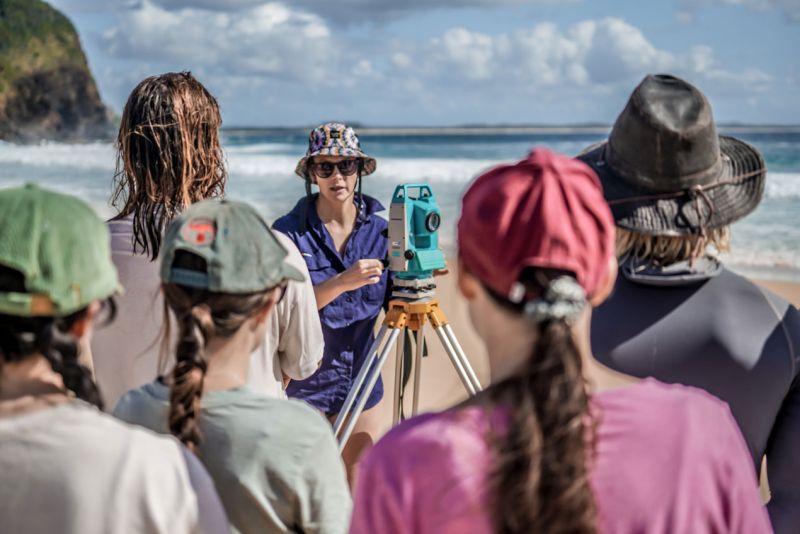 Students stand in the foreground, while focused in the background is a staff member demonstrating a surveying tool. 