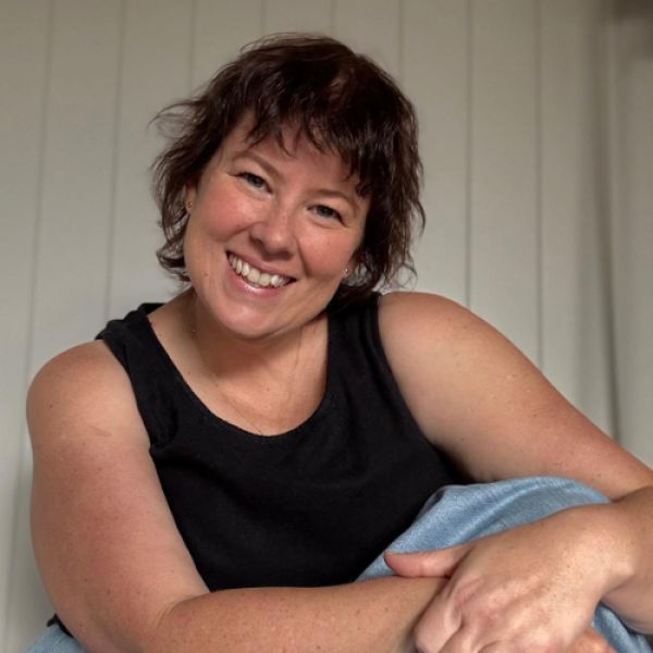 Smiling woman with short, wavy hair in a black tank top, seated against a light-colored, paneled wall. Her relaxed pose conveys warmth and friendliness.