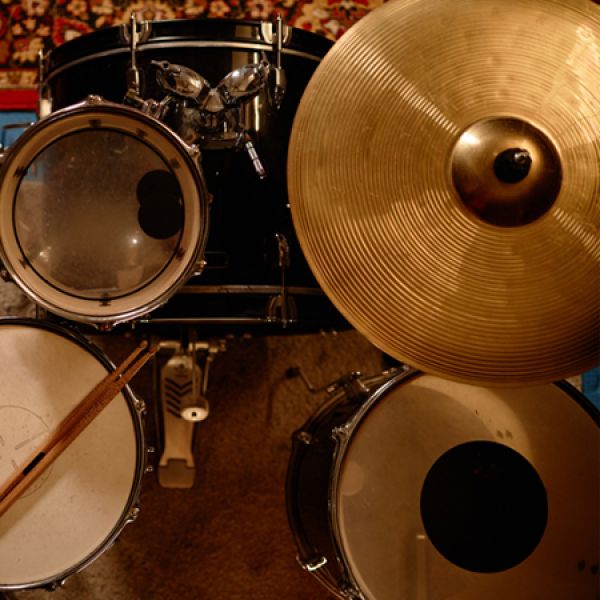 Close-up, top-down view of a drum kit with two tom drums, a snare with a drumstick resting on it, a foot pedal, and a large golden cymbal dominating the right side.