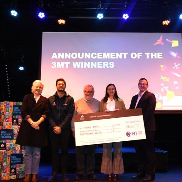 Woman holding a novelty cheque with three men and another woman standing beside her, all smiling for the camera. Social justice researcher takes out top honours at Three Minute Thesis final.