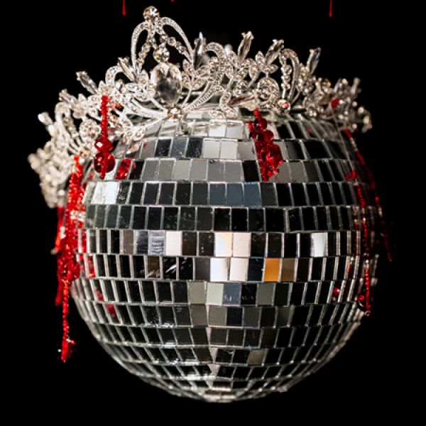 A silver mirrored disco ball topped with an ornate jeweled tiara and draped with dangling red crystal accents hangs against a black background. - Thursday 3 September 2026
