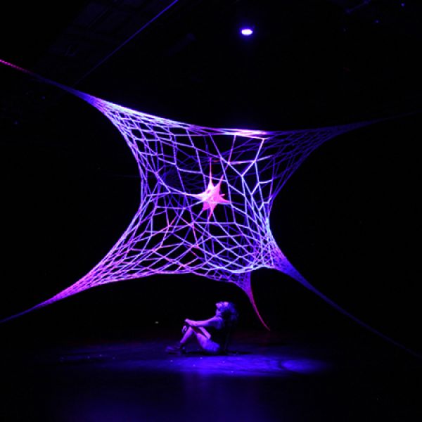 Large illuminated web-like installation in purple and blue light suspended above a seated performer in a dark gallery space.