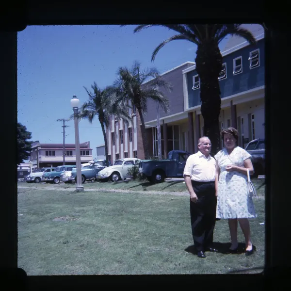 Two people stand on a grassy lawn beside palm trees, parked vintage cars and a large brick building under a clear blue sky.