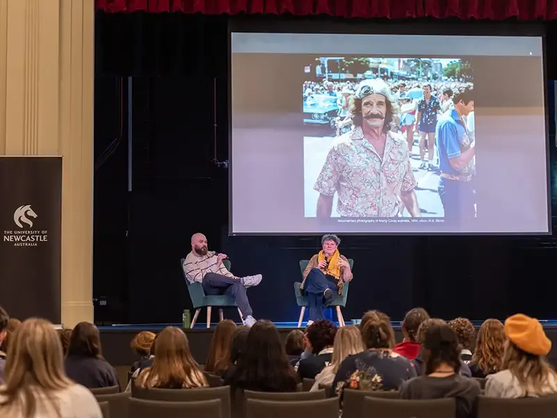 Two presenters sit on chairs on stage, presenting to a large group of students.