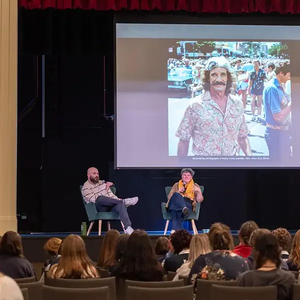 Two presenters sit on chairs on stage, presenting to a large group of students.