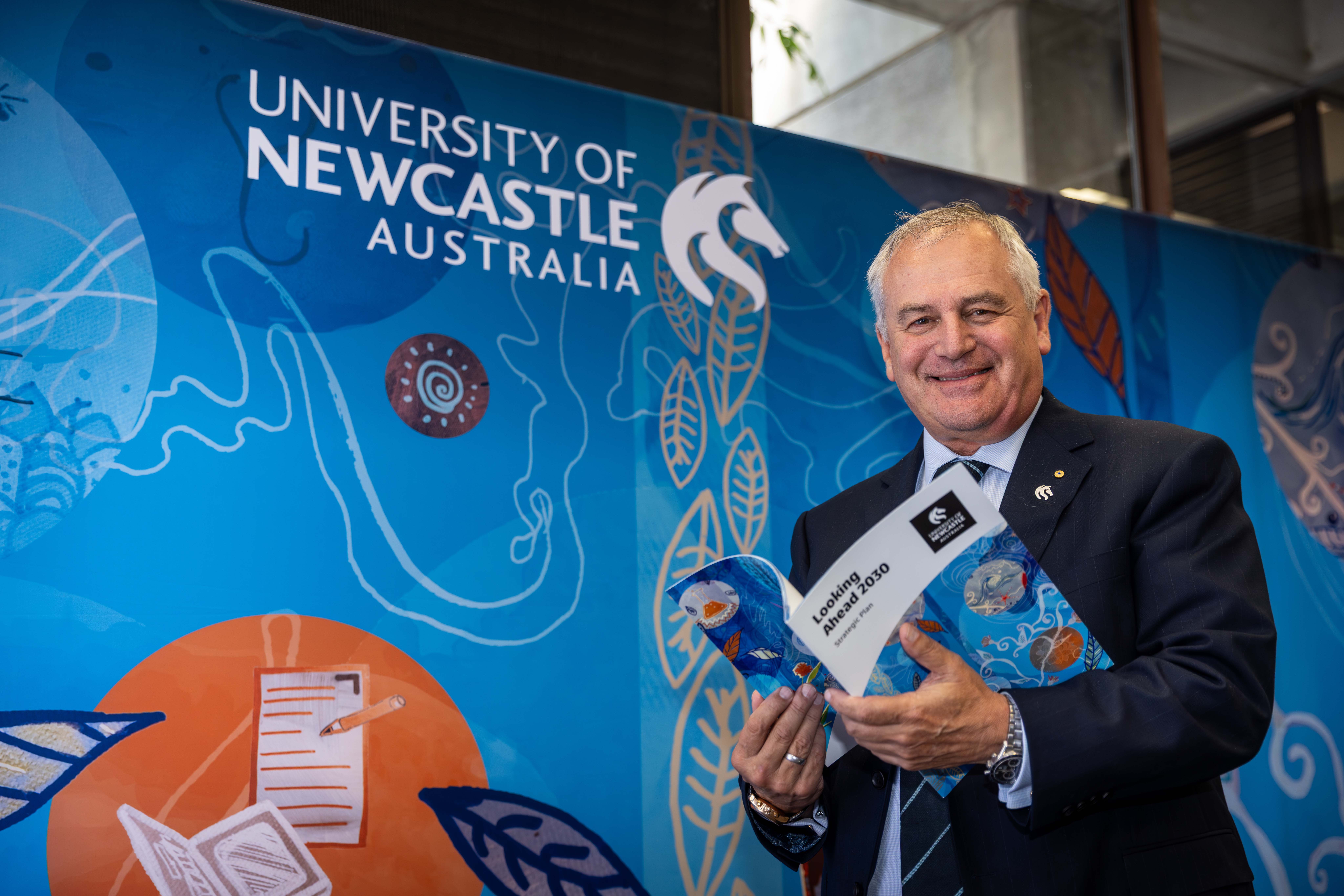 Vice-Chancellor Professor Alex Zelinsky Professor Alex Zelinsky stands looking at the camera holding a copy of the strategic plan. He is standing in front of a backdrop that has the university logo and an artwork primarliy in blue.