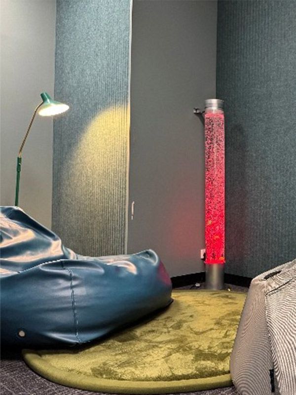 A blue bean bag sits on top of a yellow rug. An over-head light is above the bean bag and a pink lava lamp is on the right.