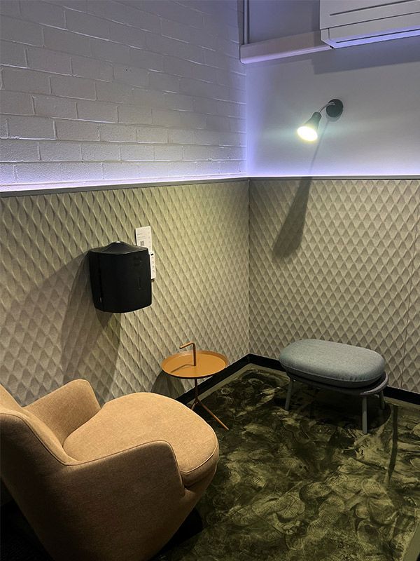 A small grey stool and brown chair is on top of a green rug. There is a small brown table in between the stool and chair.