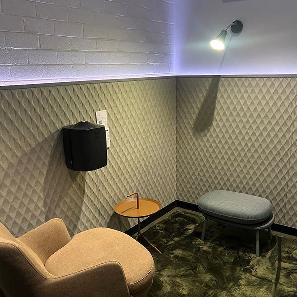 A small grey stool and brown chair is on top of a green rug. There is a small brown table in between the stool and chair.