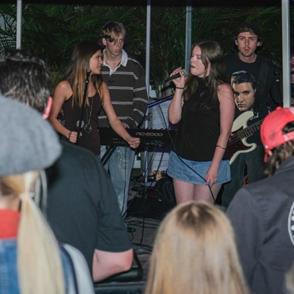 A band performs in front of an audience, with a vocalist singing into a microphone while other musicians play keyboard and guitar in the background.