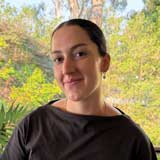 Head-and-shoulders portrait of a person wearing a dark top and hoop earrings, photographed outdoors with trees and foliage in the background.