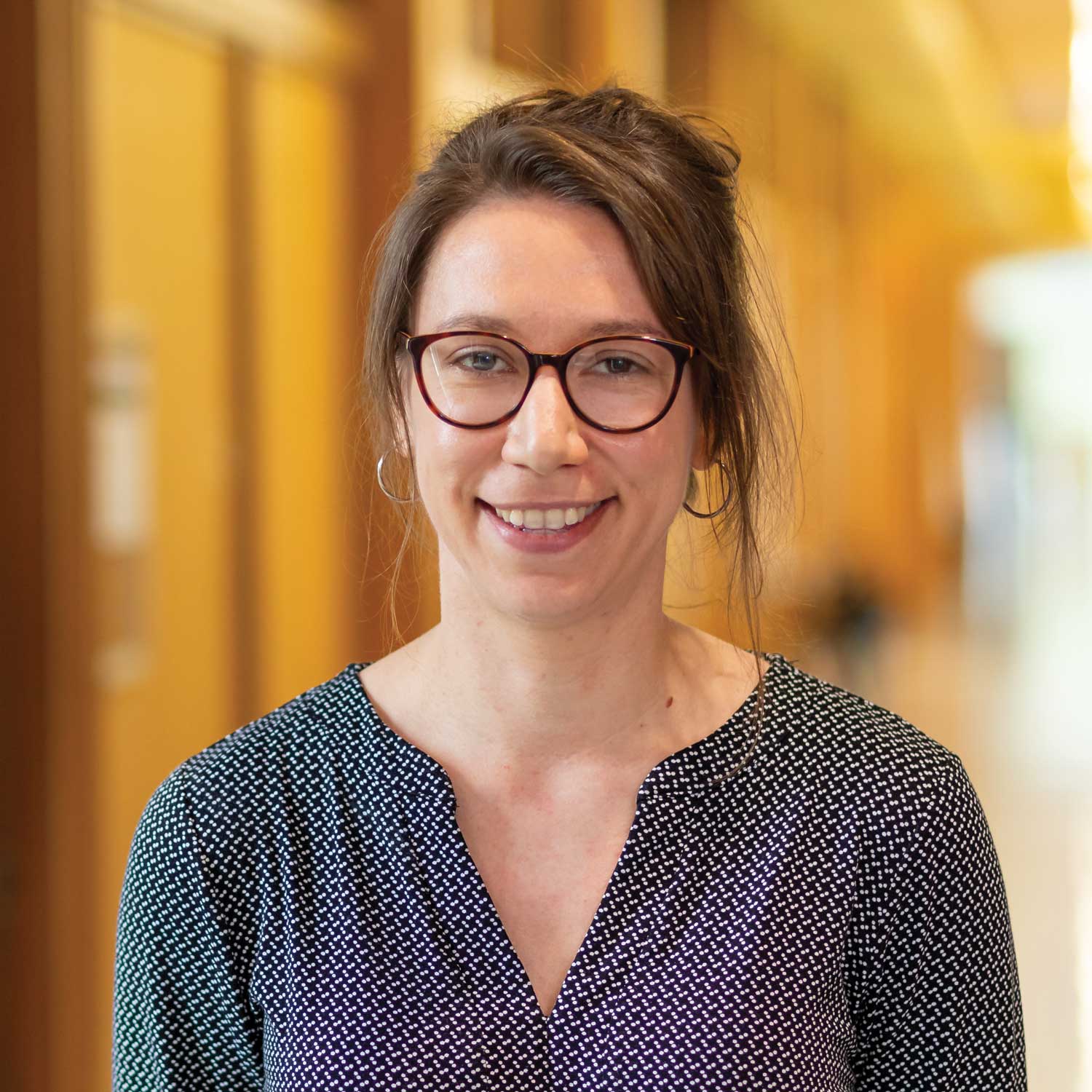 Head-and-shoulders portrait of a person wearing glasses and a dark patterned blouse, standing indoors in a softly lit corridor. The background is blurred, with warm-toned timber walls and architectural details creating a professional university setting.