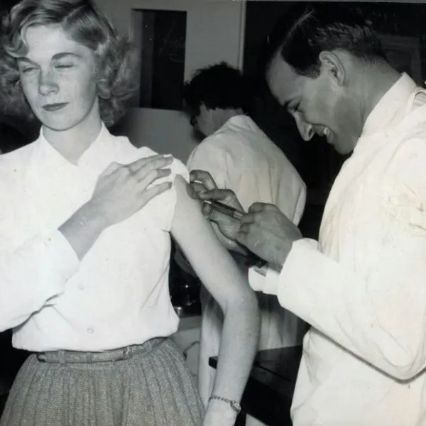 Black-and-white historical photograph of a healthcare worker administering an injection into a person's upper arm while another person in a white coat stands in the background.