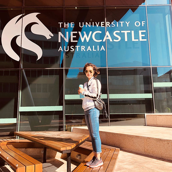 A person standing on a table holding a coffee in front of a University of Newcastle logo printed on a building