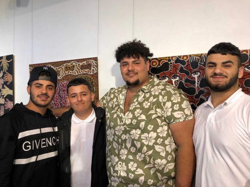 Four young men with their arms around each other standing in front of Aboriginal paintings