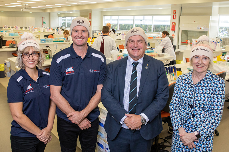 Four people stand in a lab looking at the camera.They are all wearing blue and Mark Hughes Foundation beanies