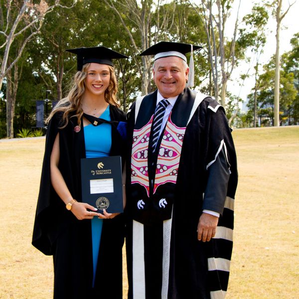 Jessica Haugh in a graduation gown stands next to the Vice-Chancellor wearing his official gown. They are standing on a grassy area and there are people in the background behind them. The sky is blue