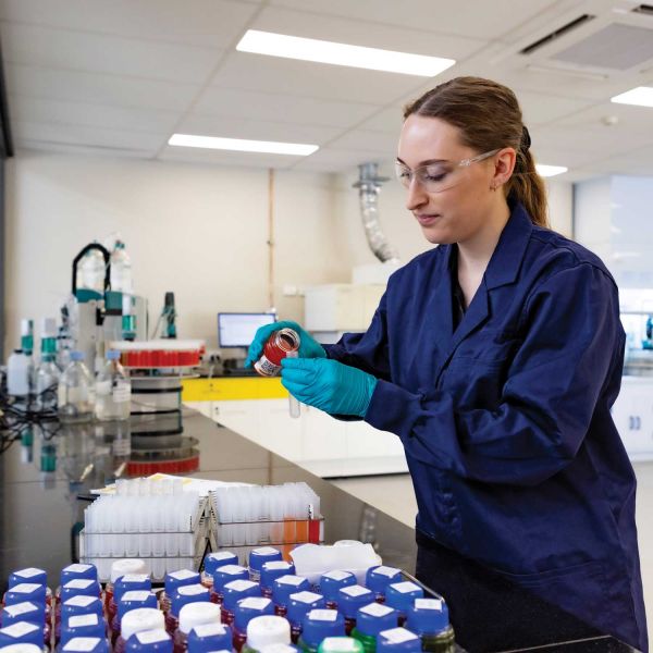 A female scientist in a laboratory pouring liquid into a test tube. The scientist is wearing a lab coat, safety glasses and gloves