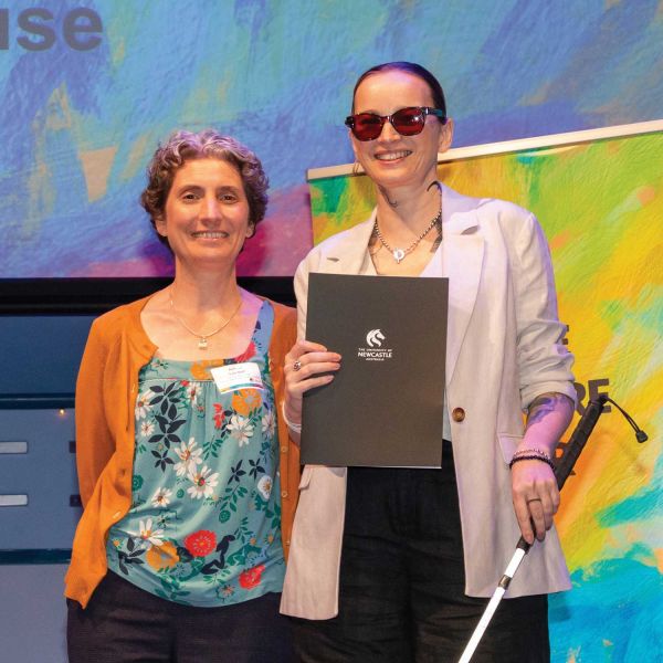 Two women on stage at a presentation with a colourful artwork in the background. One woman is holding an award. 