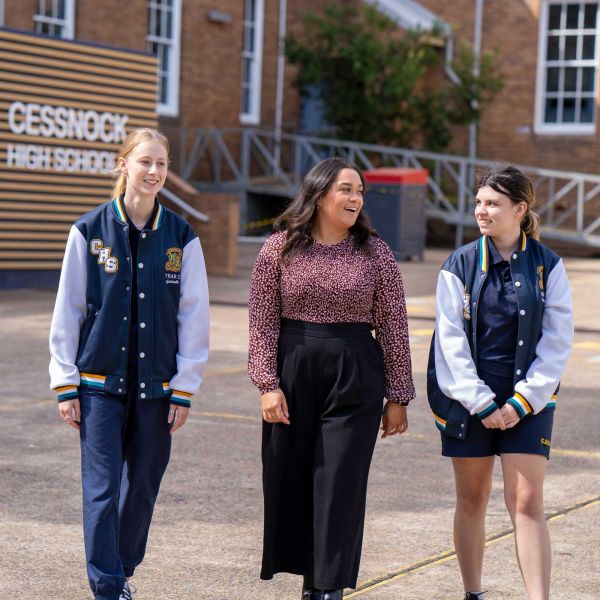 Cessnock High School teacher Kelsey (centre) walks with two Year 12 students. Cessnock High School is in the background of the image.. 75 years of innovation in teacher education.