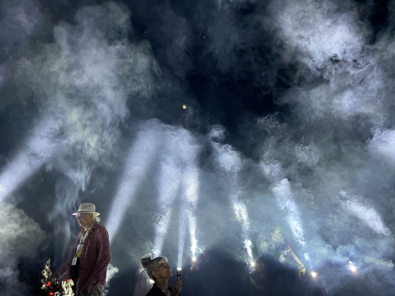 People sitting in a circle using torches pointed up at the sky to create a tipi shape using light.