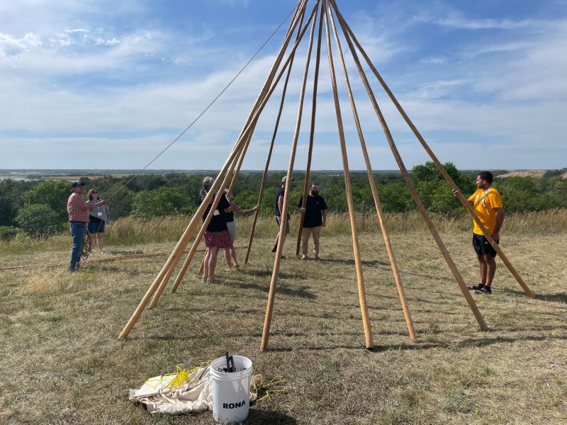 Ten or twelve tall sticks arranged in a circle tipi shape. There are six people around it holding ropes and preparing to finish the tipi.