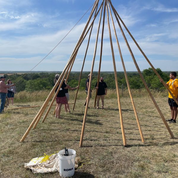 Ten or twelve tall sticks arranged in a circle tipi shape. There are six people around it holding ropes and preparing to finish the tipi.