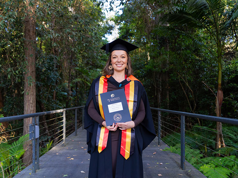 Kiah Carmody O'Toole stands in front of a bushland backdrop proudly holding her degree and wearing cap and gown