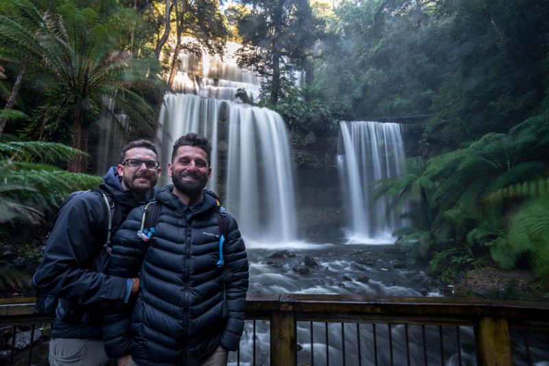 Sonny and his husband smiling in front of a waterfall, surrounded by lush greenery and sunlight filtering through the trees.
