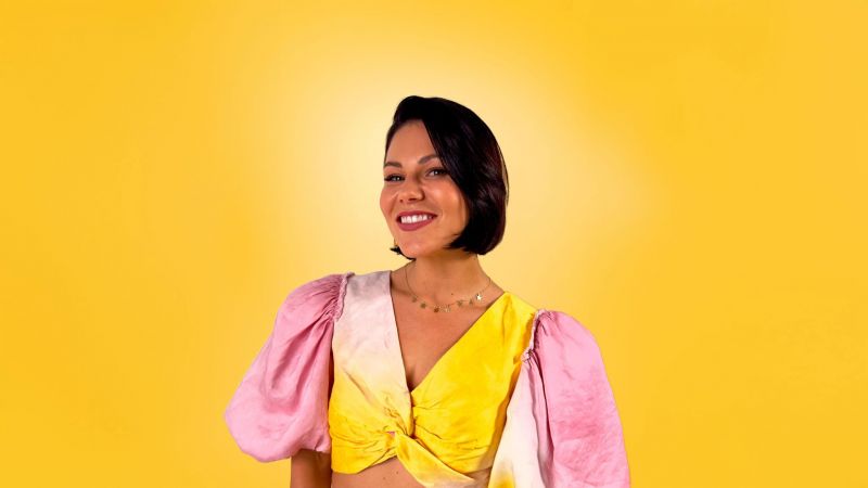 Storm smiling, dressed in a yellow and pink outfit, standing against a yellow background.