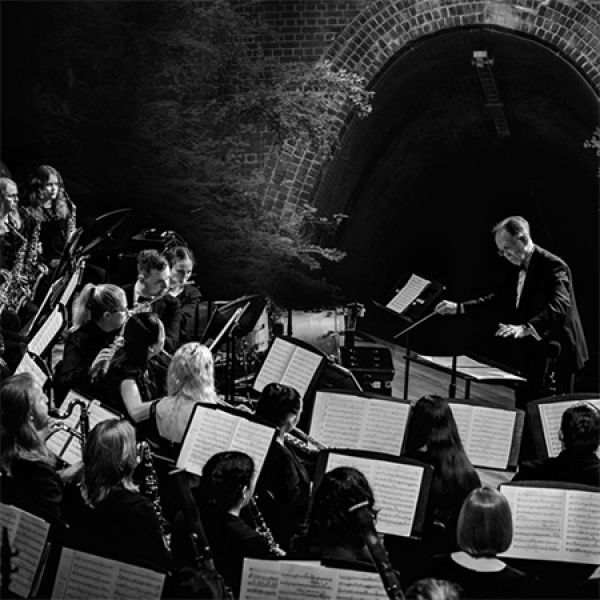 Black and white photo of an orchestra performing in front of a tunnel, led by a conductor.