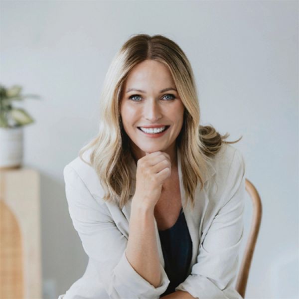 A person with shoulder-length blonde hair in a light blazer and dark top sits on a chair with one hand on their chin.