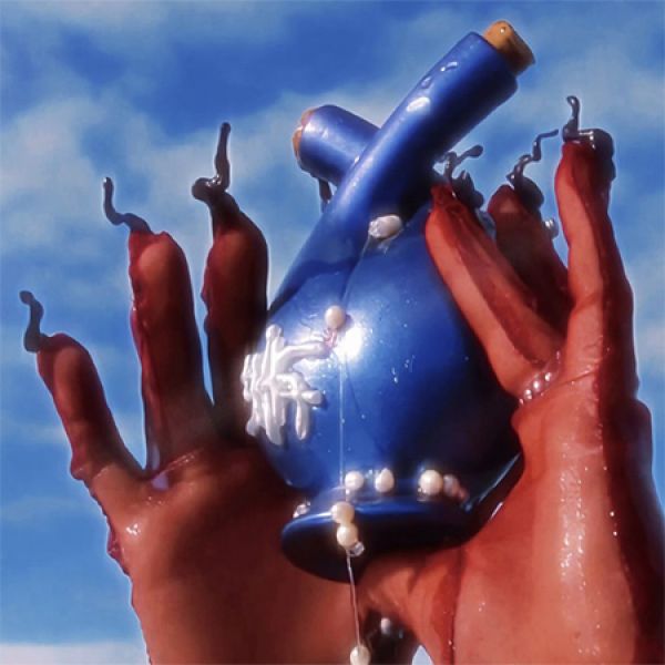 Bio Assembly Exhibition Two hands covered in a red substance hold a blue ceramic object with white decorations against a clear blue sky.