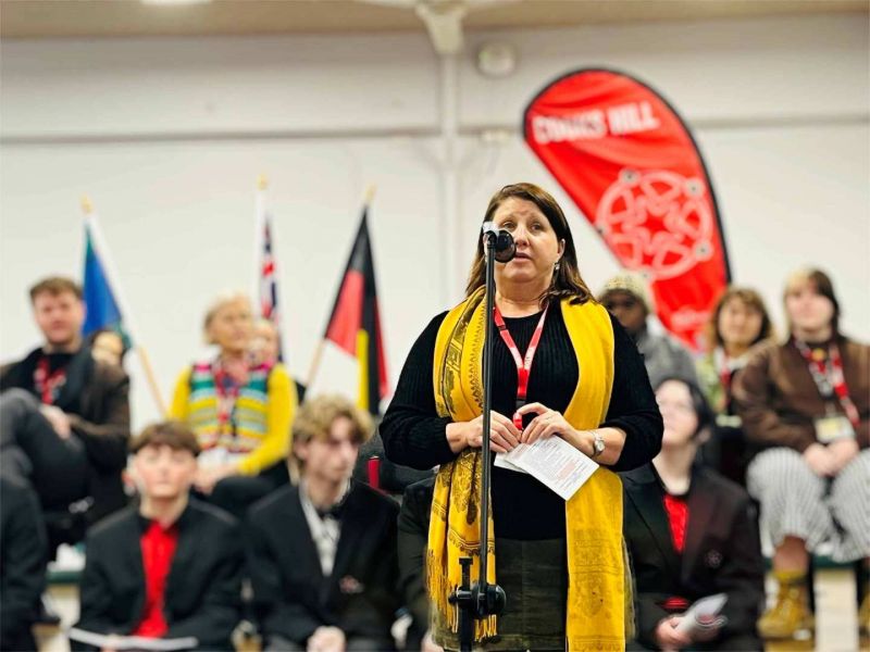 A woman with a yellow scarf standing in front of a microphone with people in chairs seated behind her