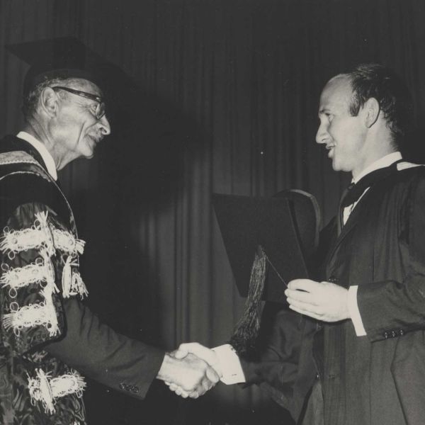 Image of a graduate with their cap off shaking the hand of a Professor in robes and a cap. It is black and white and from 1966.. Celebrating 60 years of Community Support.