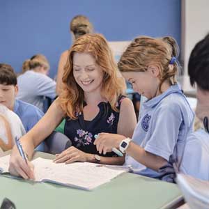A teacher is holding a pen and smiling with a school student standing next to them in a classroom surrounded by students