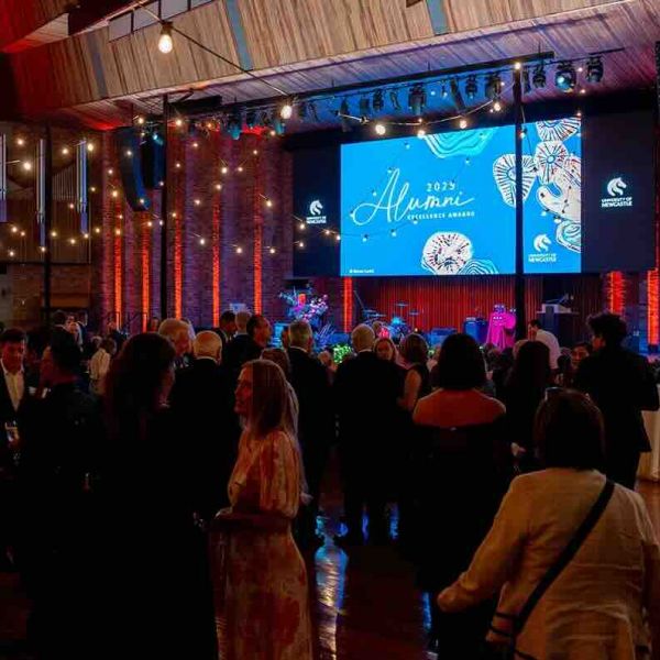 A crowded Great Hall with a blue screen on stage with the title of 2025 Alumni Excellence Awards. University of Newcastle commemorates graduate excellence .