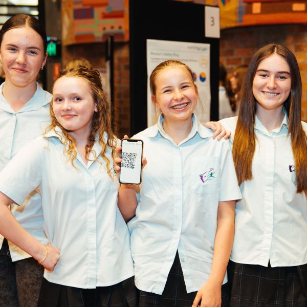 A group of four girls smiling at the camera. One holds a phone, demonstrating a prototype. A research poster is displayed behind the students.